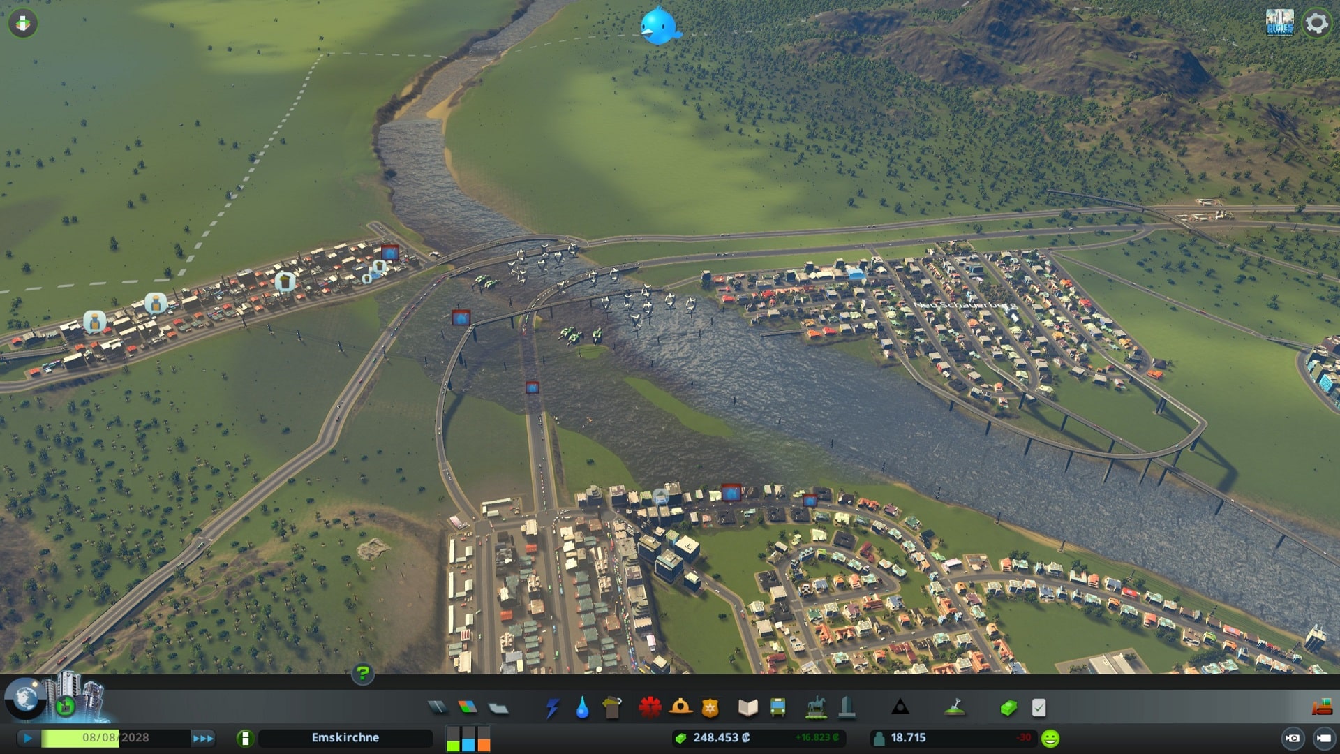 Dargestellt ist ein Screenshot aus der Städtebau-Simulation Cities: Skylines. Zu sehen ist eine kleine Stadt, die sich an einen Fluss schmiegt. Das Wasser des Flusses tritt an vielen Stellen über die Ufer und hat Teile der Stadt unter Wasser gesetzt. 