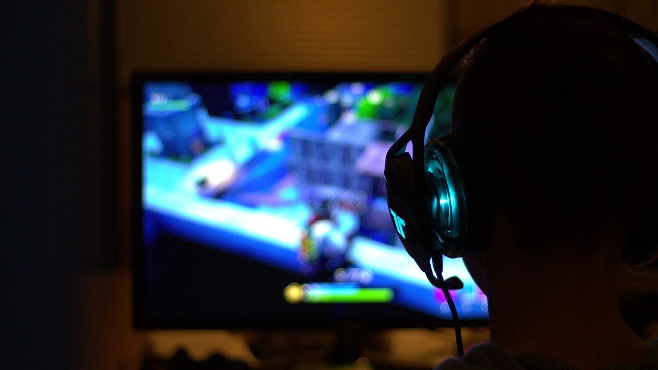 Im Vordergrund ist die Silhouette eines Kopfes samt Headset zu sehen. Der dargestellte Spieler blickt auf einen Bildschirm im Hintergrund. Auf diesem ist schemenhaft ein Spiel (Fortnite) zu sehen, welches der Spieler gerade Spielt. 