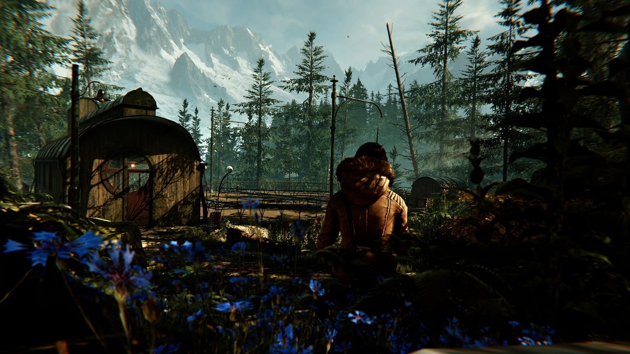 Screenshot aus dem Spiel Syberia: The World Before. Zu sehen ist die junge Kate Walker, die in einer idyllischen Berglandschaft sitzt und auf eine kleine Bahnstation blickt. 